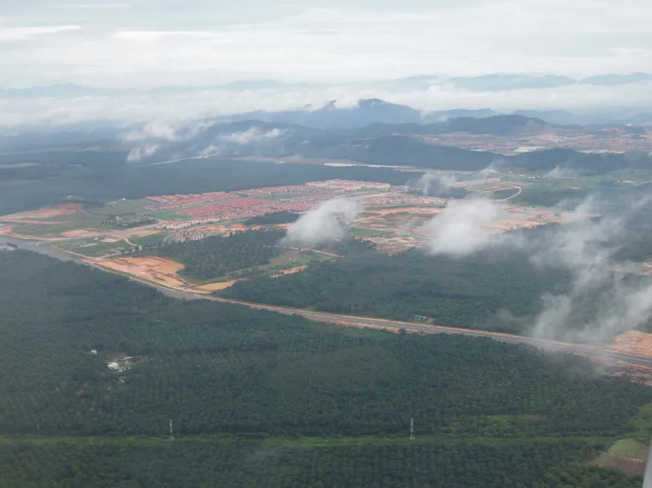 Aerial photo of cleared, reddish-brown land beside a highway, with a housing development, forested hills and low cloud in the distance.